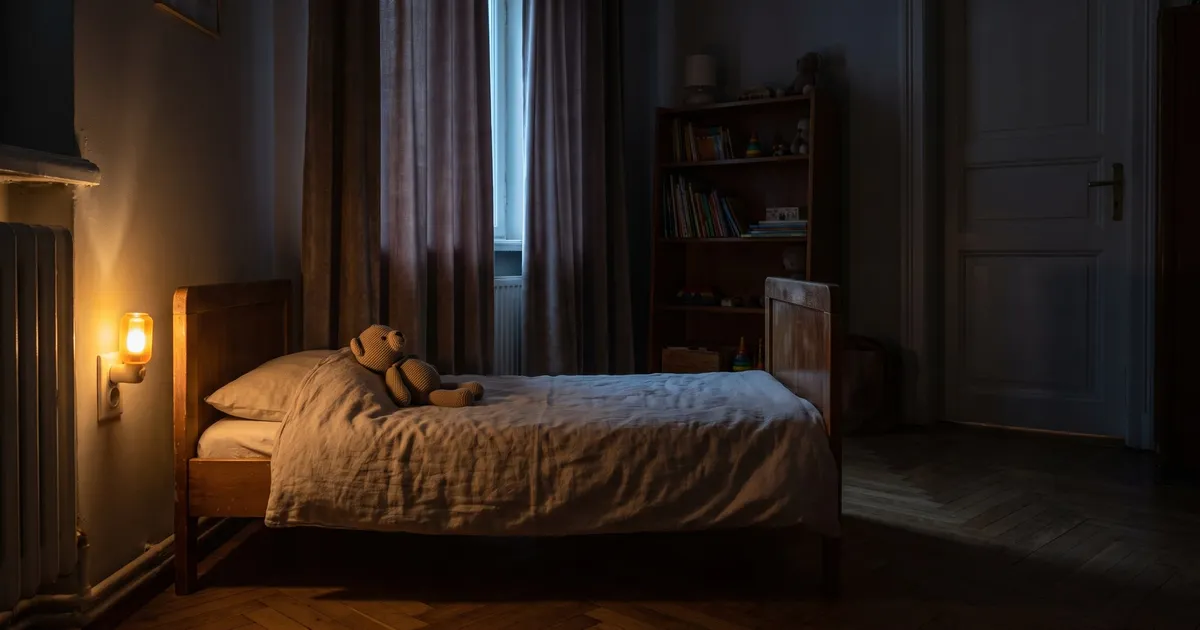 Symbolbild (KI-generiert): dunkles Kinderzimmer, ein warmes Steckdosen-Nachtlicht neben einem niedrigen Holzbett